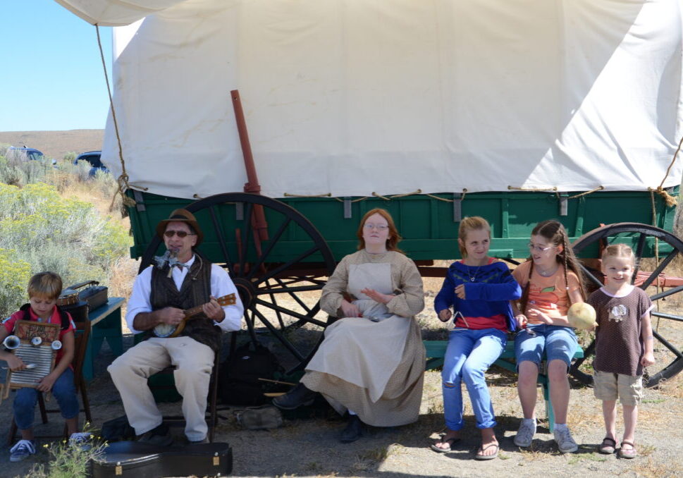 pioneer and children playing vintage musical instruments in front of a covered wagon